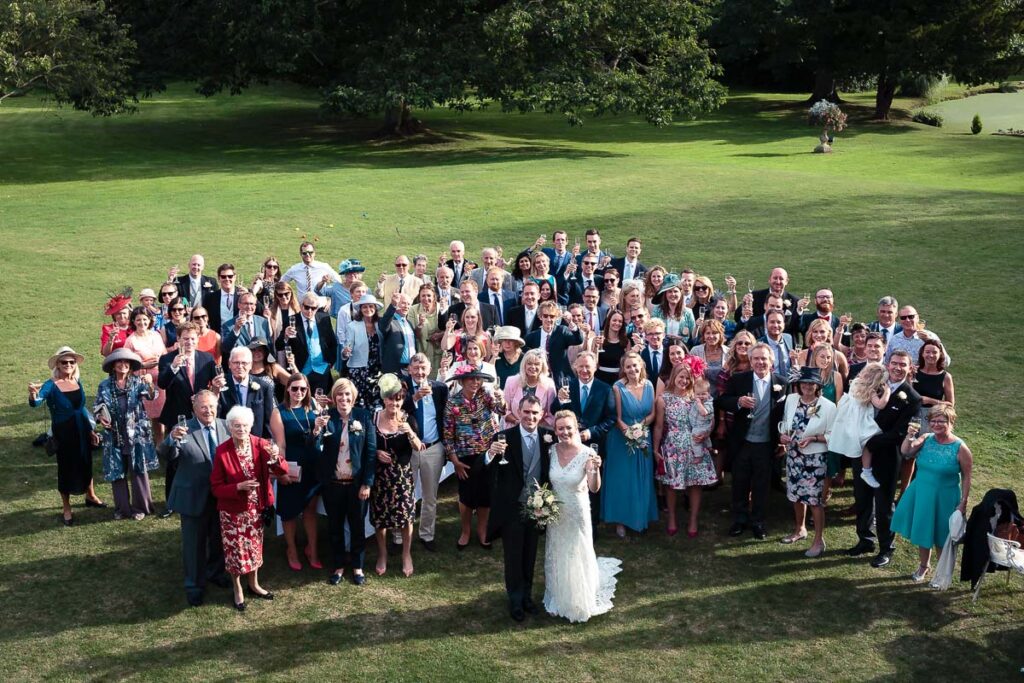 A large wedding group photograph taken outdoors on a sunny day in a park with green grass and mature trees in the background.