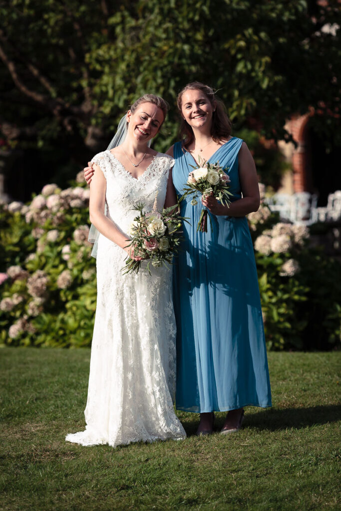 Two women in wedding attire standing together on a lawn, holding flower bouquets, smiling at the camera with flowering shrubs behind them.