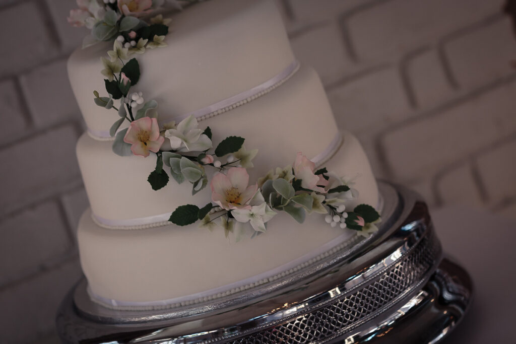 A three-tiered white wedding cake decorated with pale pink flowers and green foliage, displayed on a silver cake stand.