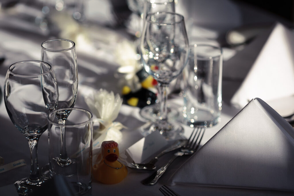 A formally set dining table with empty glasses, white napkins, cutlery, and a small decorative duck, bathed in sunlight.