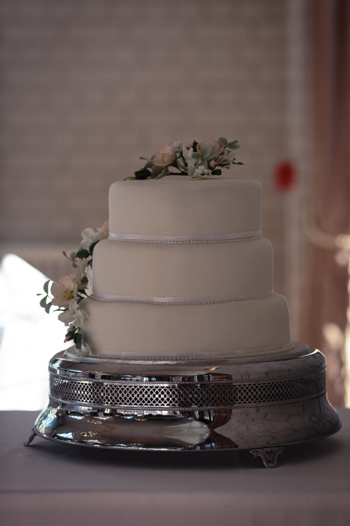 A three-tiered white wedding cake with pearl beading, topped with succulents and flowers, sits on an ornate silver cake stand.