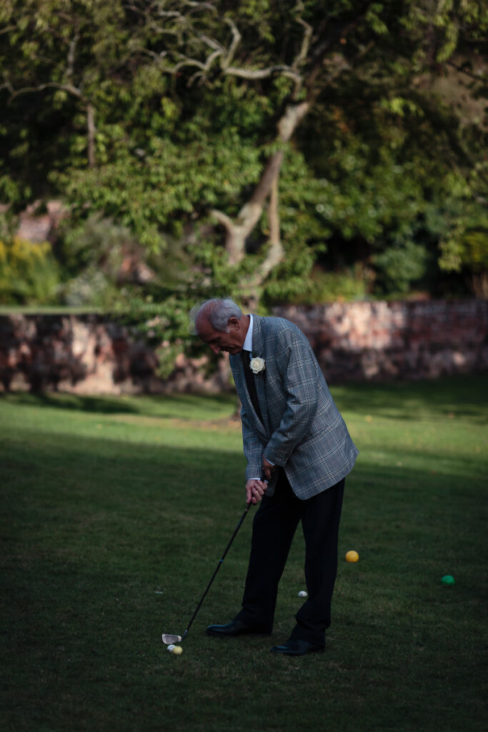An elderly man in a blue checked jacket plays croquet on a lawn, preparing to strike a white ball with his mallet.