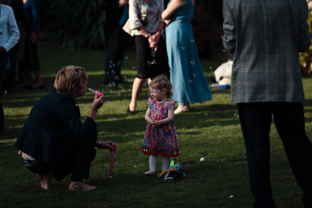 A young child in a floral dress blows bubbles while an adult crouches beside her on a grass lawn, surrounded by formally dressed guests at an outdoor event.