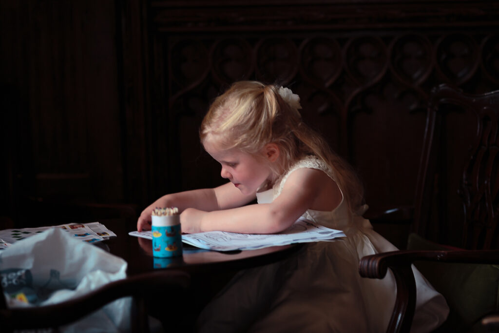 A young child with blonde hair sits at a dark table, drawing or writing on paper with a blue container nearby.