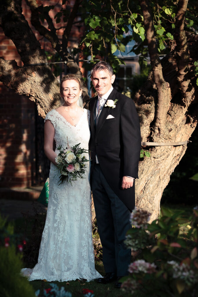 A bride and groom smile at the camera, standing together beneath a large tree. She wears a lace wedding dress and holds a bouquet; he wears a dark suit.