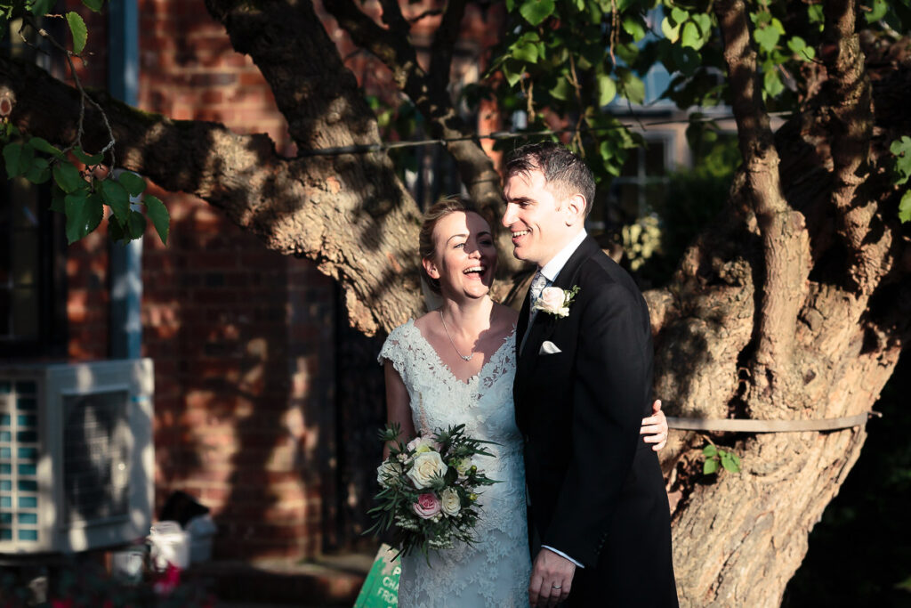 A bride and groom stand together beneath a large tree, smiling at each other. The bride holds a bouquet and wears a lace dress; the groom wears a dark suit.
