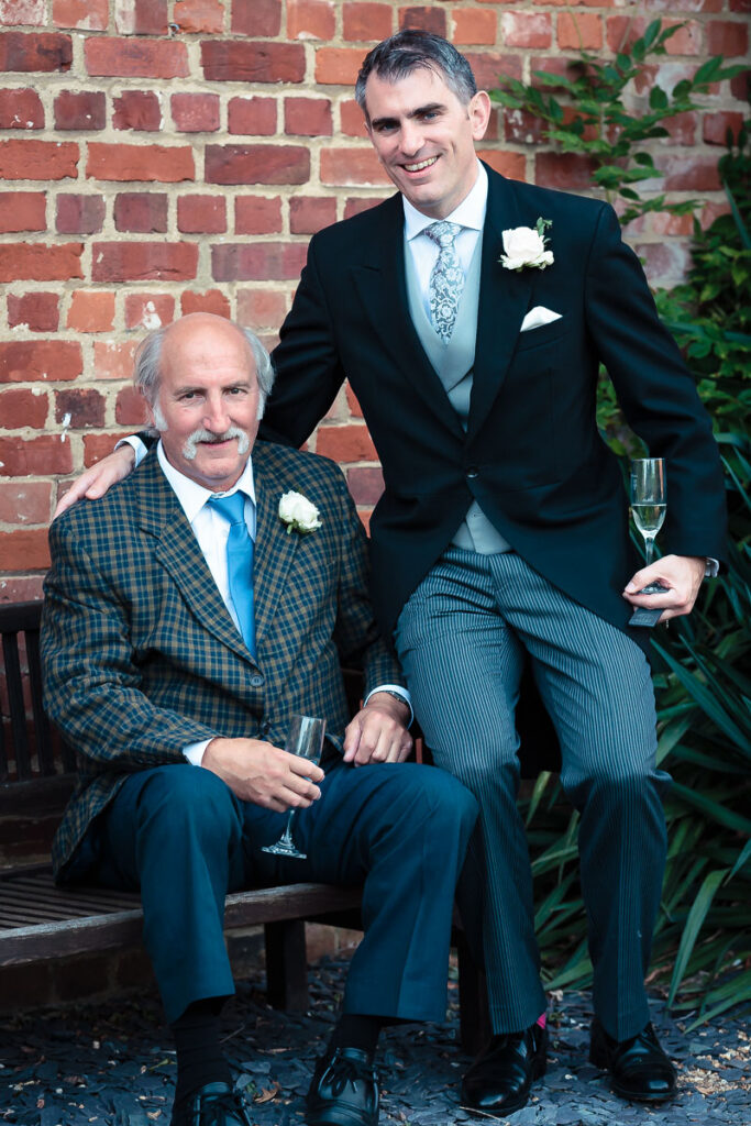Two men in formal wedding attire pose together against a red brick wall. The older man sits while the younger man stands beside him, both holding champagne glasses and wearing buttonholes.