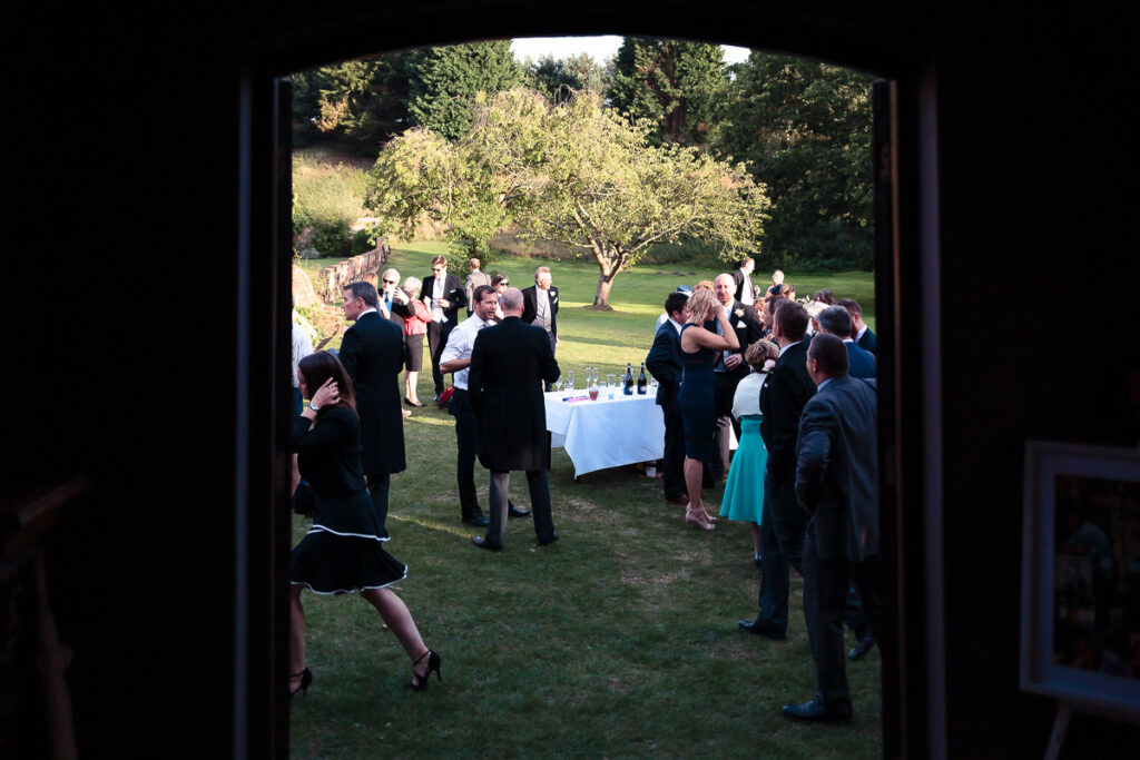 A formal garden gathering viewed through open doors. Guests in dark clothing mingle on a lawn beside a white-clothed table, with trees and green fields beyond.