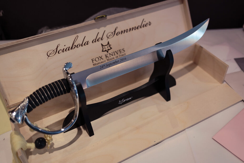 A decorative knife displayed on a wooden stand. The blade is engraved Fox Knives and dated 24th September 2016. The handle features an ornate silver guard and black grip.