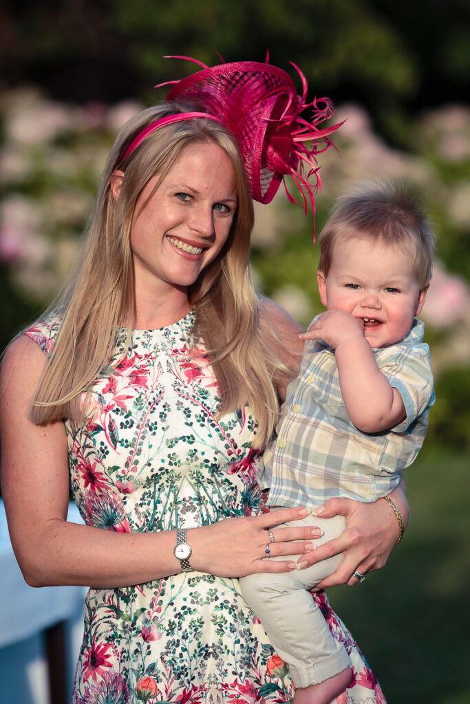 A woman in a floral dress and pink fascinator holds a smiling toddler in a checkered shirt, both outdoors on a sunny day.