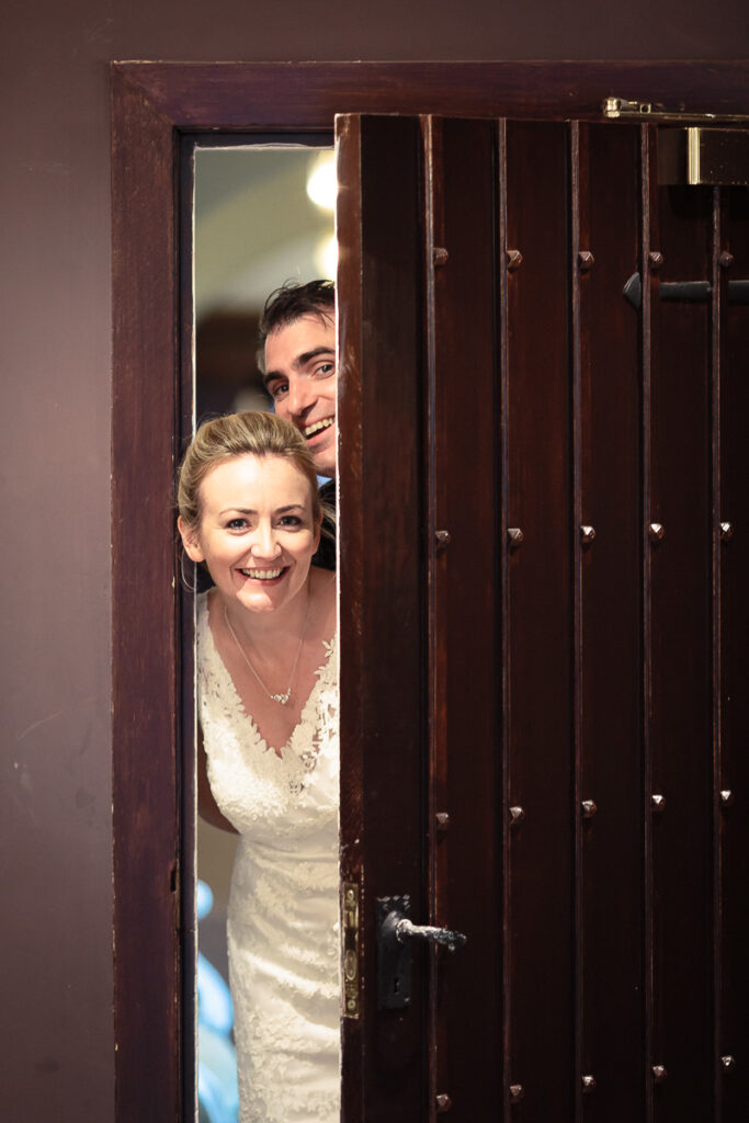 A couple peering through a dark wooden door with vertical slats, both smiling at the camera. The woman wears a white lace dress.