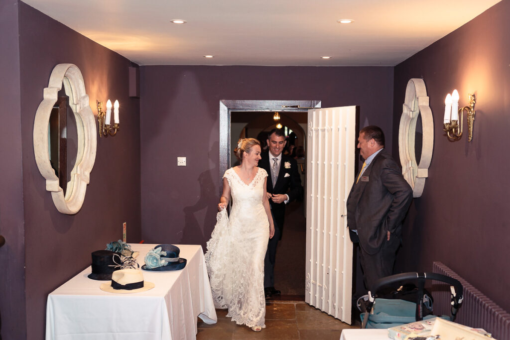 A bride and groom enter a purple-walled room while a man in a suit greets them. Decorative mirrors and candelabras line the walls.