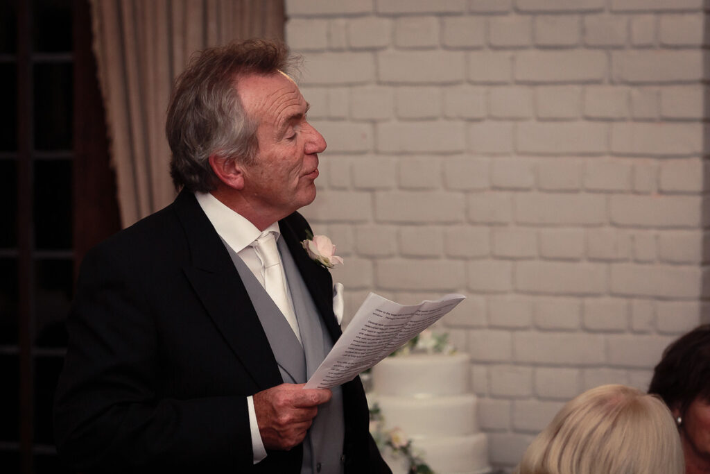 A man in formal attire holds a document, speaking at what appears to be a wedding reception indoors.