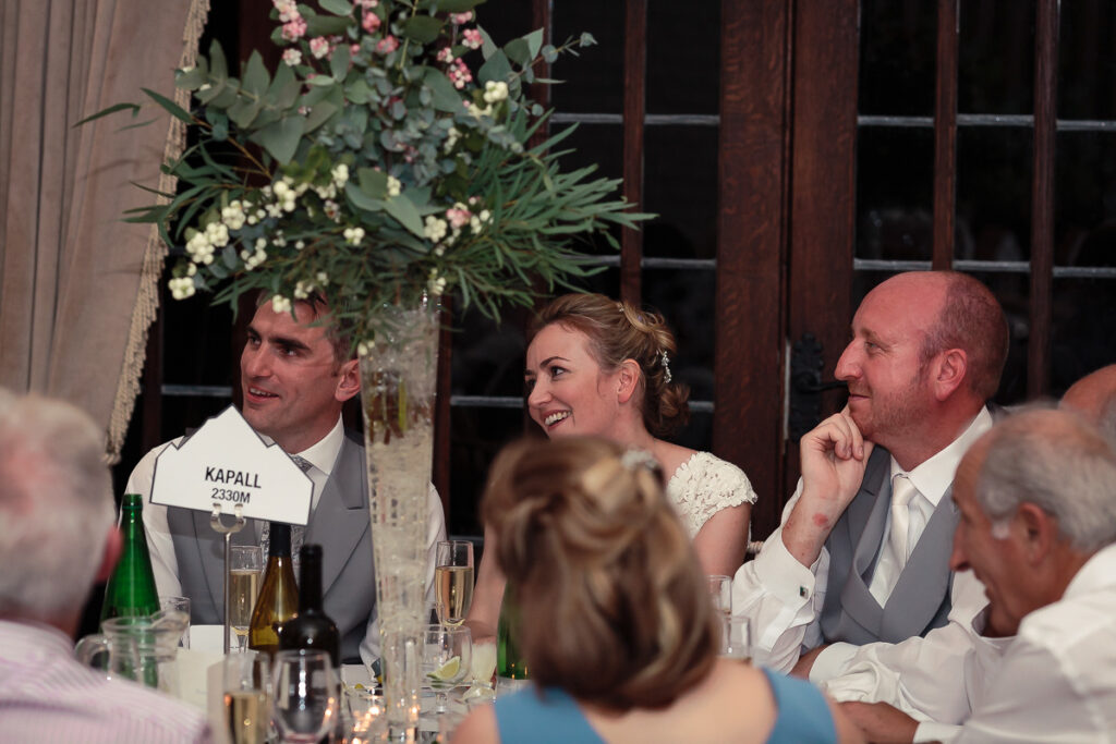 Wedding reception table with bride and groom seated among guests, elegant floral centerpiece overhead, wine glasses and bottles visible.