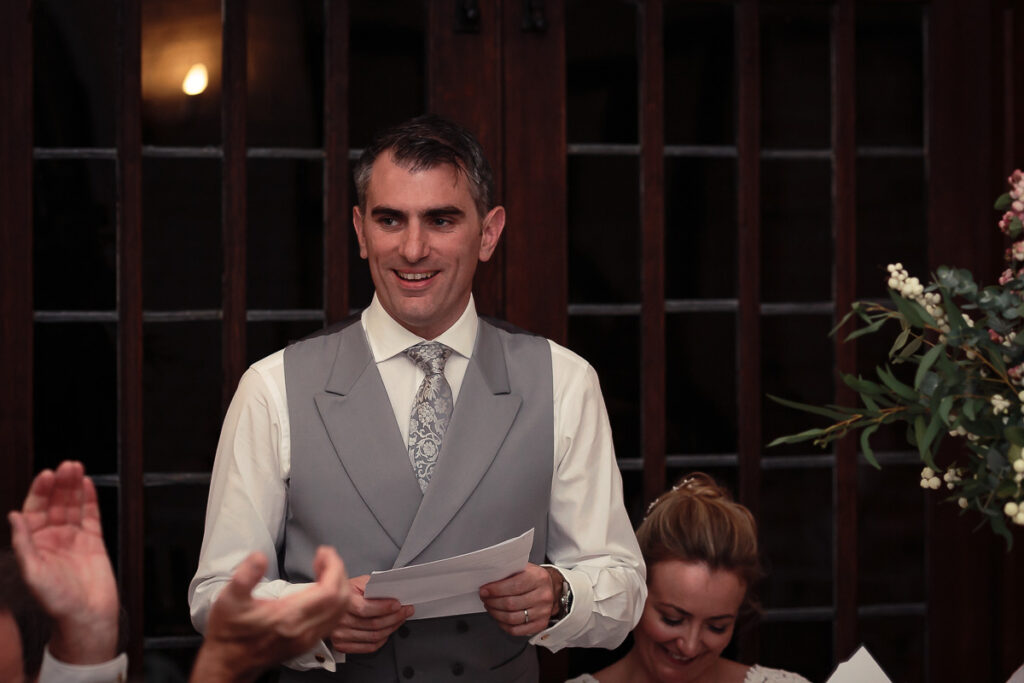A man in a grey waistcoat and white shirt holds papers while speaking at an indoor event, smiling at the camera.
