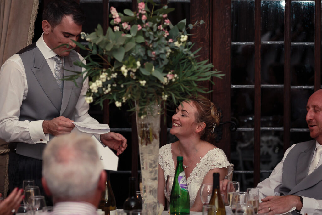 A man in waistcoat gives a speech at a formal dinner while a smiling woman in a lace dress listens.