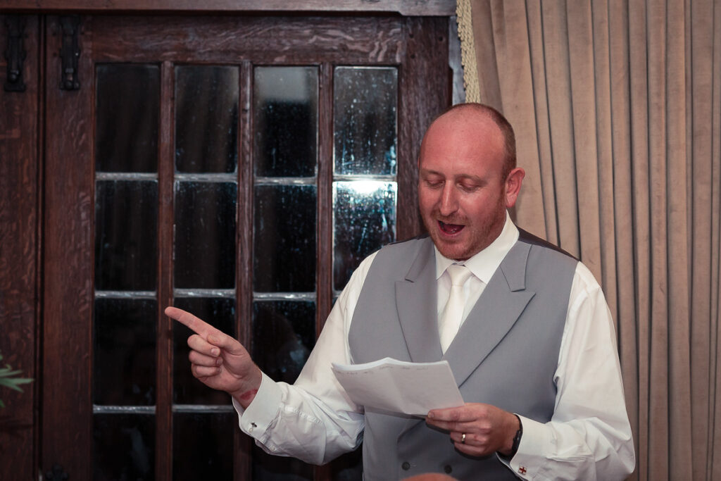 A man in a grey waistcoat and white shirt holds papers and gestures while speaking indoors, with wooden doors behind him.
