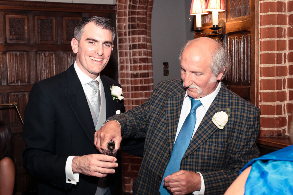 Two men in formal attire with boutonnières at an indoor event. The man on the left wears a black suit and holds a cigar. The man on the right wears a checkered jacket and blue tie. Brick walls and wooden paneling visible in the background.