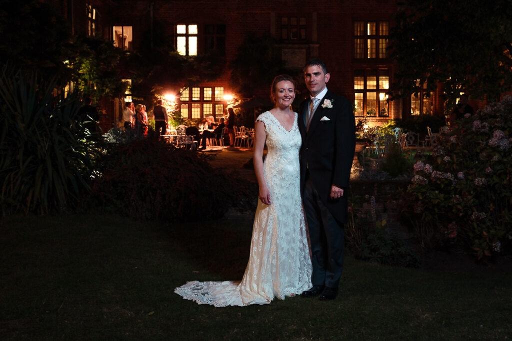 A bride and groom stand together in a garden at night, with lit windows of a brick building behind them and flowering plants on either side.