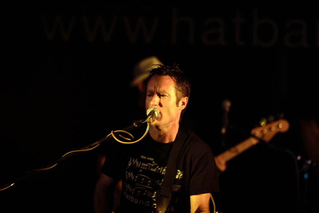 A male musician performs on stage at night, singing into a microphone whilst wearing a black graphic t-shirt. A bassist is visible in the background.