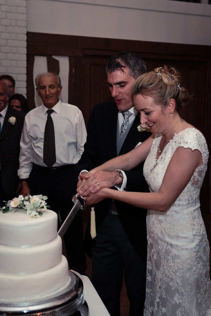 A bride and groom cut their wedding cake together at their reception, surrounded by guests in formal attire.