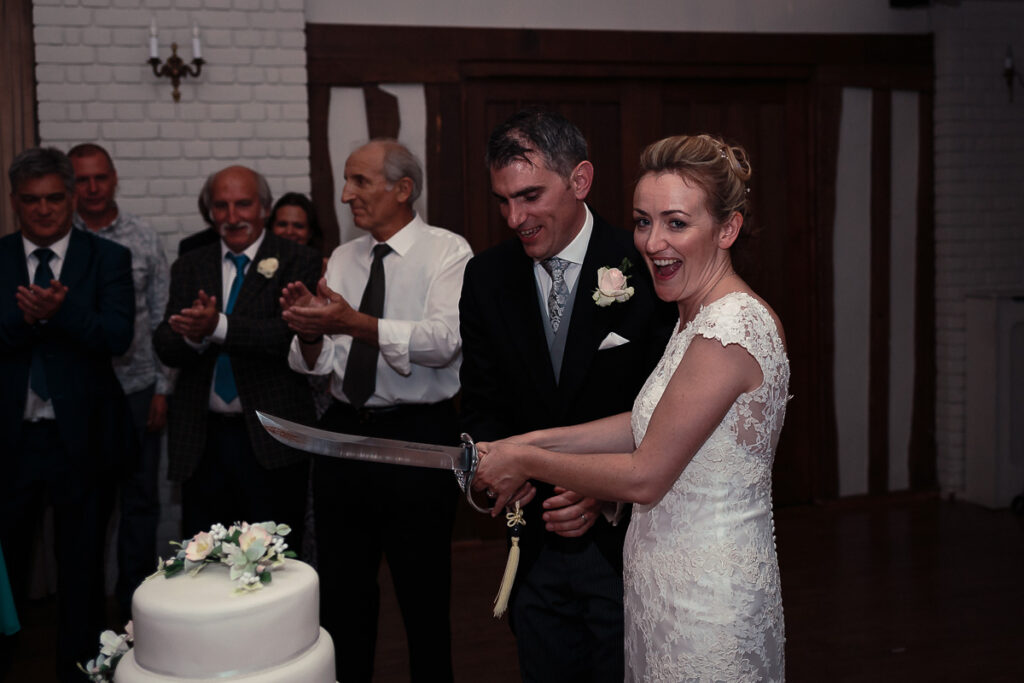 A bride and groom cut their wedding cake together with a sword while guests applaud in the background.