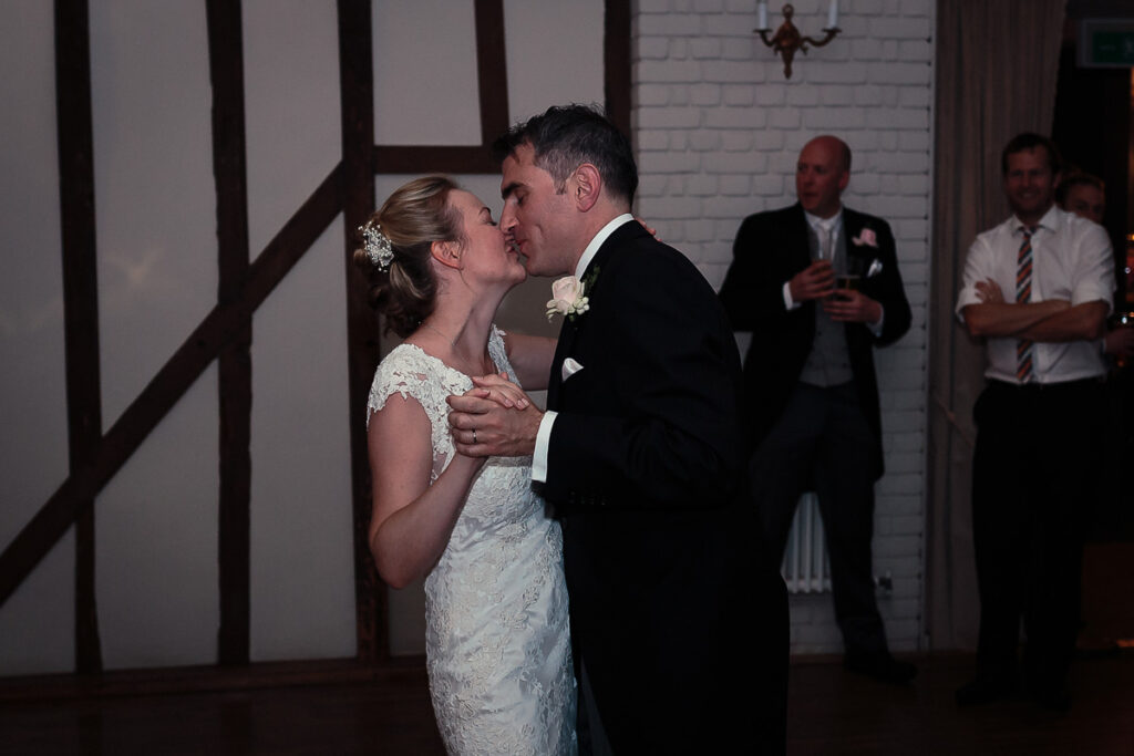 A bride and groom kiss during their first dance at a wedding reception, watched by guests in formal attire indoors.