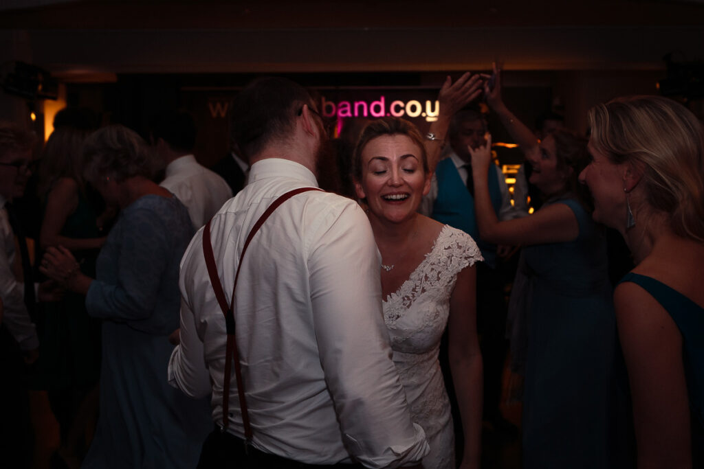 A bride and groom dancing at a wedding reception. The bride wears a white lace dress and smiles brightly. The groom wears a white shirt with a burgundy tie, facing away from the camera. Other guests surround them on a dark dance floor.