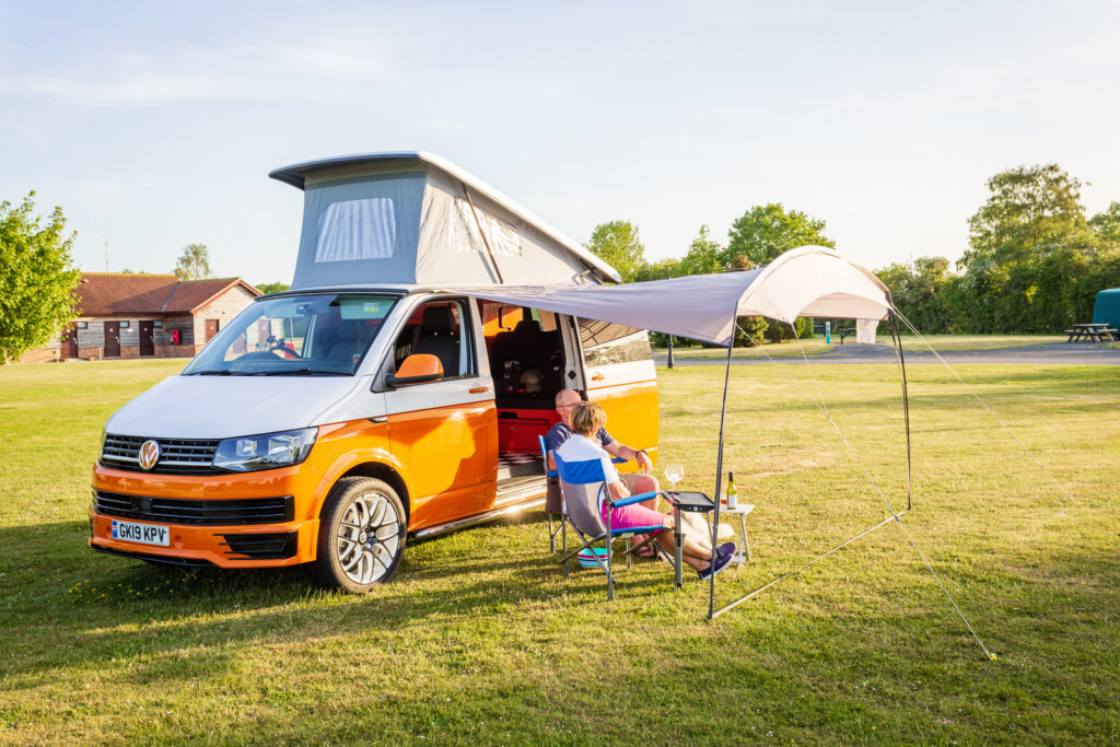 Orange and white VW campervan with its roof raised and awning out on a grassy campsite