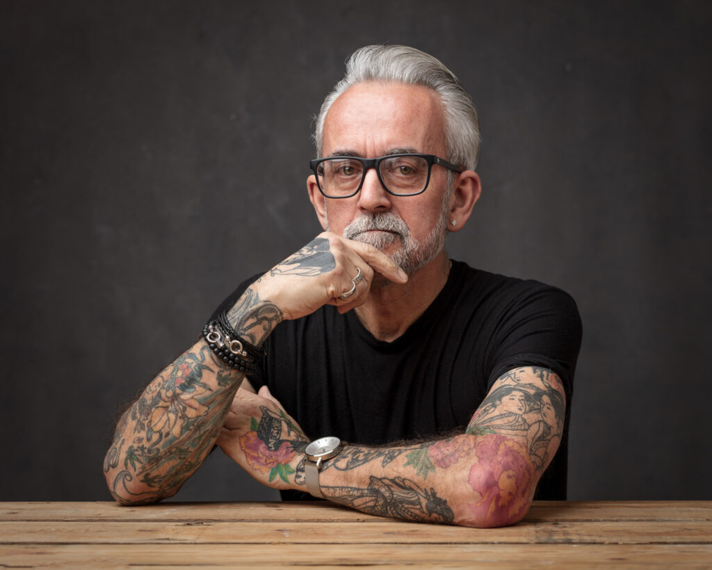 Personal branding portrait of a tattooed man with glasses leaning on a wooden table