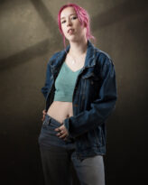 Woman with pink hair in a denim jacket and green crop top against a studio backdrop