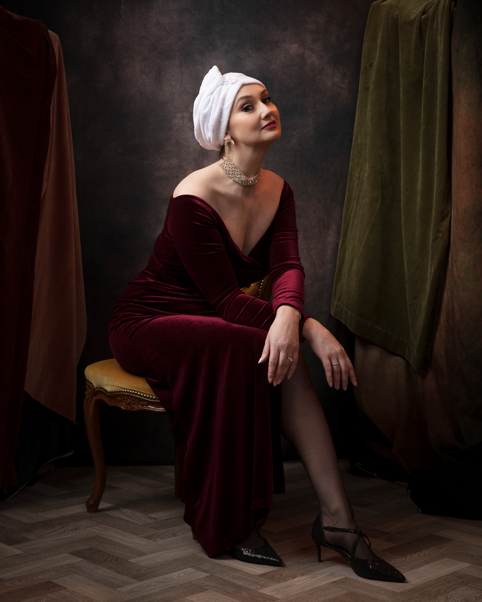 Studio portrait of a model in a burgundy velvet dress and white head wrap, seated in a painted set