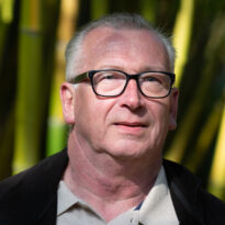 Portrait of an older man with grey hair and black glasses against a bamboo backdrop