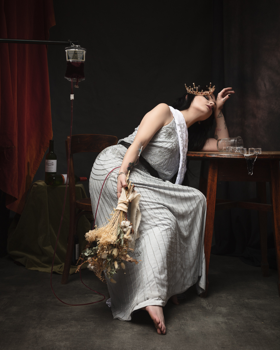 Woman in crown with IV and bouquet, tired pose.
