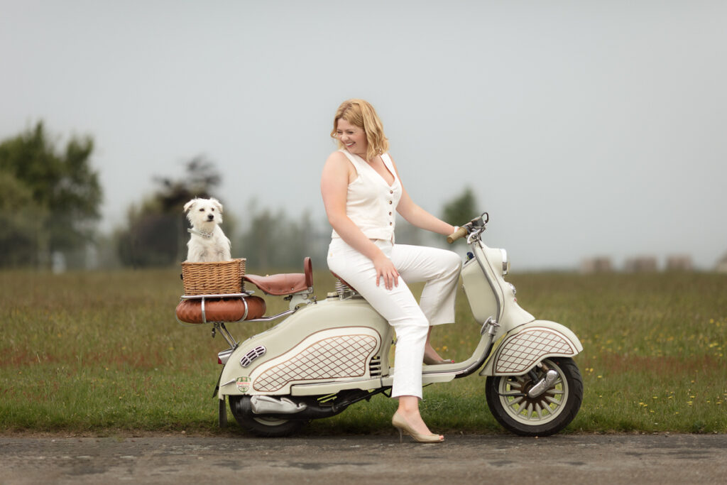 Woman in white sitting on a cream scooter in a field, with a small white dog in a wicker basket, portraying a serene outdoor setting.
