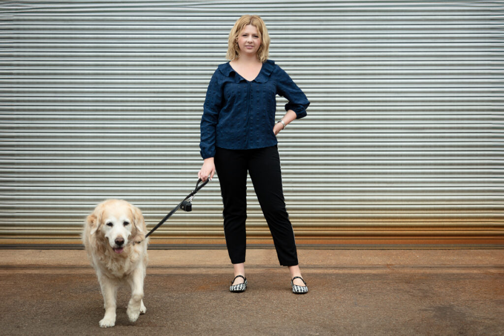 Woman in blue blouse and black trousers standing with a golden retriever against a corrugated metal background.