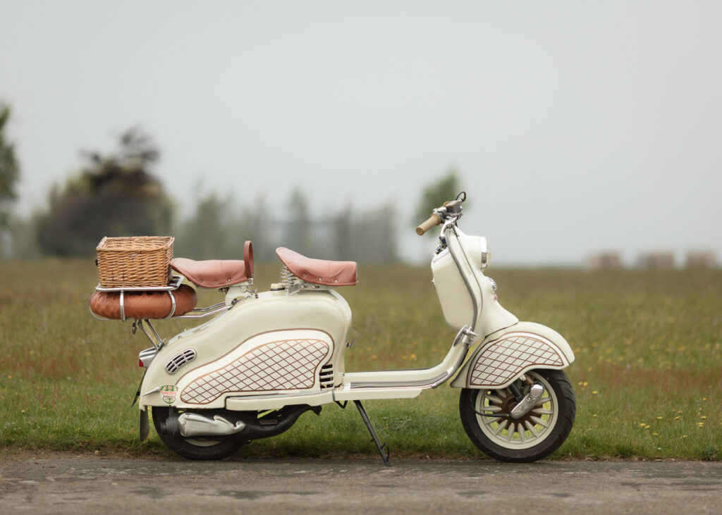 Vintage cream-colored scooter with a wicker basket and leather accessories parked in a grassy field.