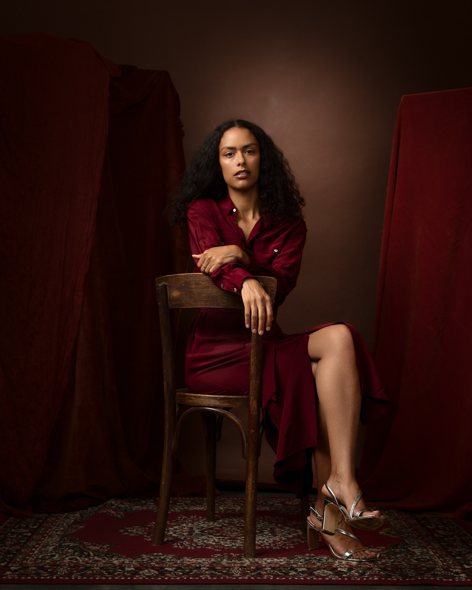 Woman in burgundy shirt seated on wooden chair, dramatic red background