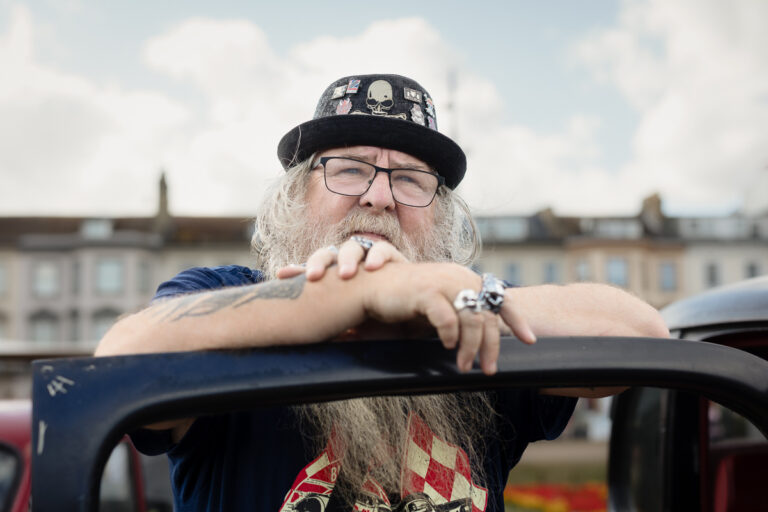 Bearded man wearing skull cap and glasses leaning on car
