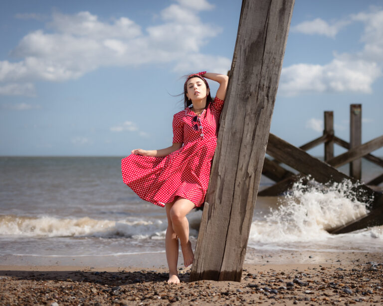Woman in red polka dot dress posing by wooden post on beach