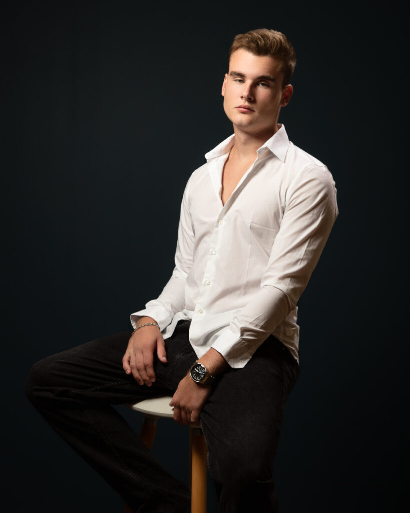 Man sitting on stool wearing white shirt and jeans.