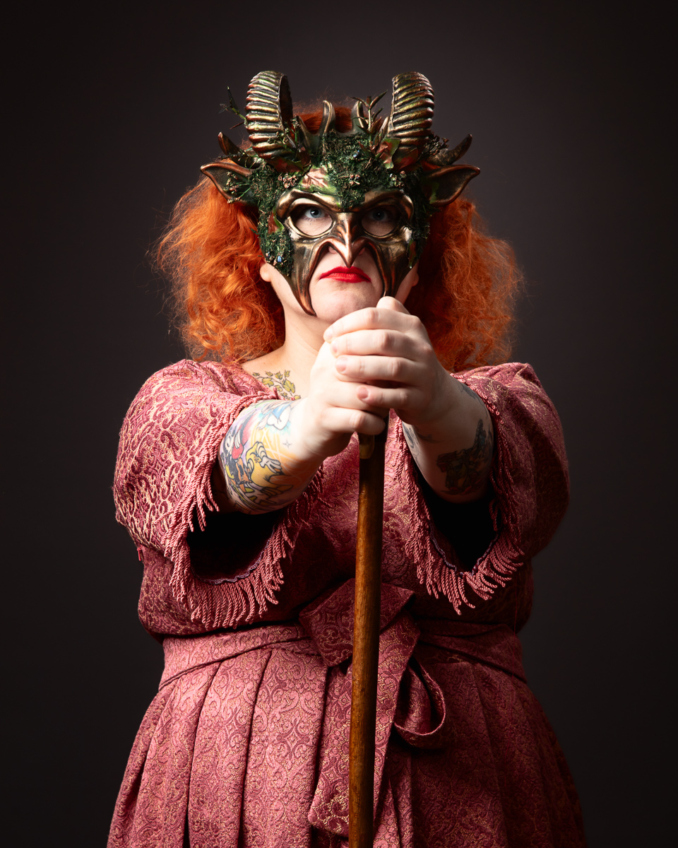 Performer in elaborate demon mask, red hair, pink costume with staff