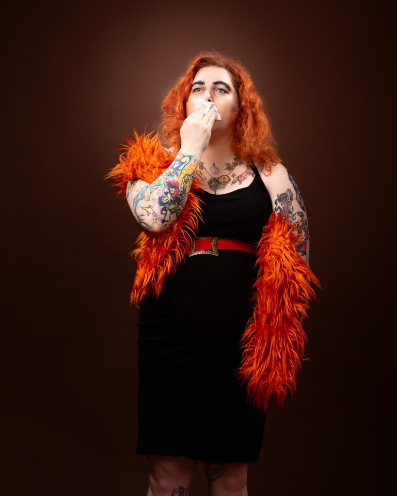 Person with tattoos and orange boa blowing nose.
