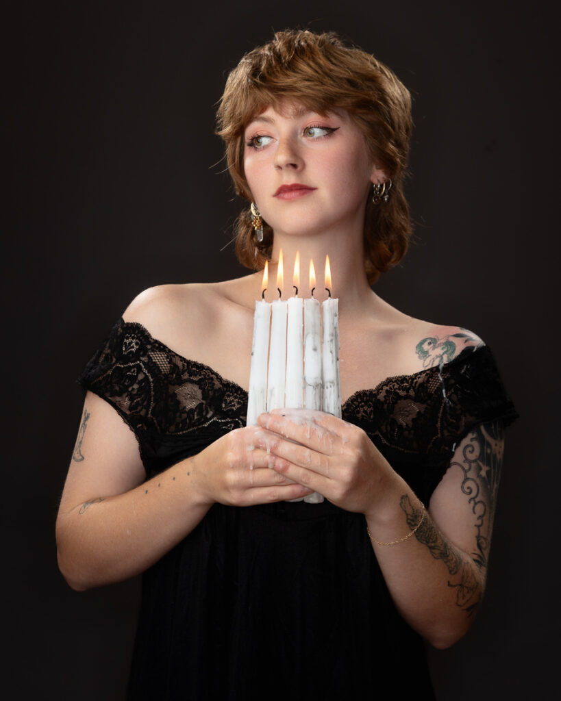 Person holding lit white candles, black dress, tattoos.