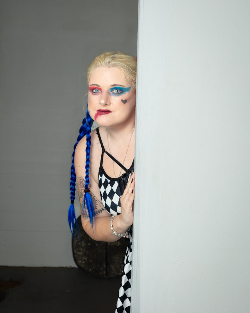 Woman with blue braids and colourful eye makeup peering around wall