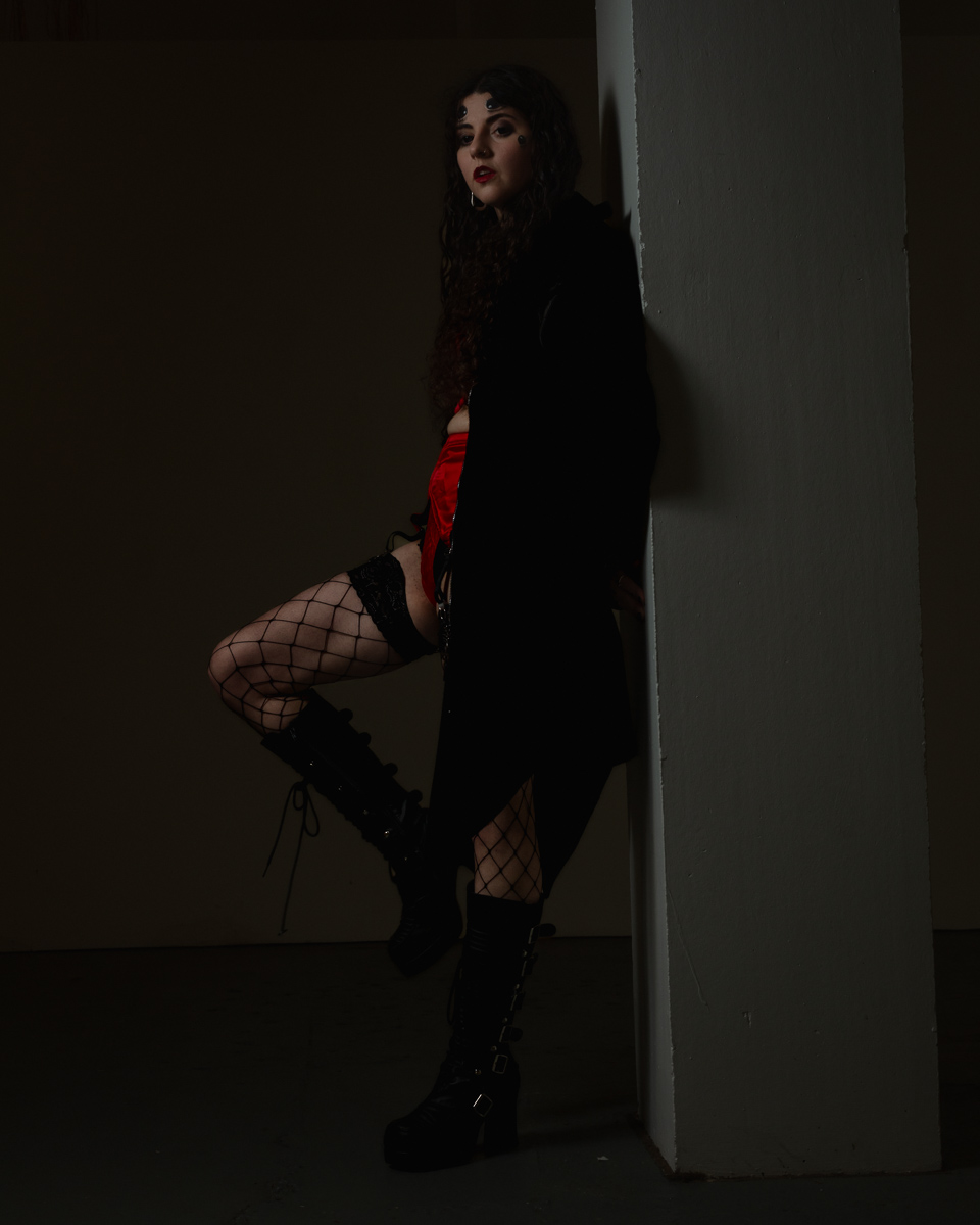 Woman in black coat and fishnet stockings poses against wall