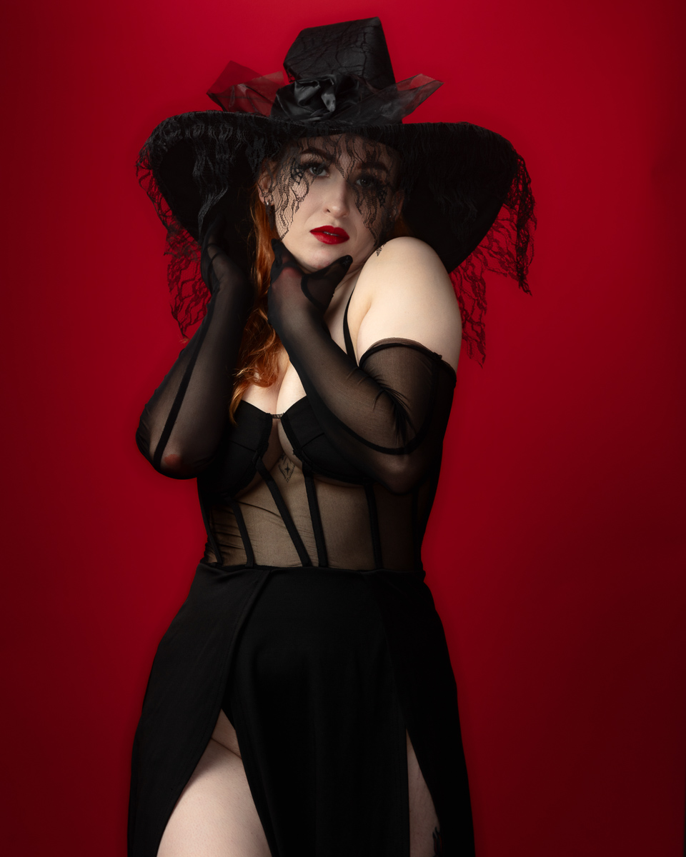 Woman in black gothic outfit with wide-brimmed hat against red background