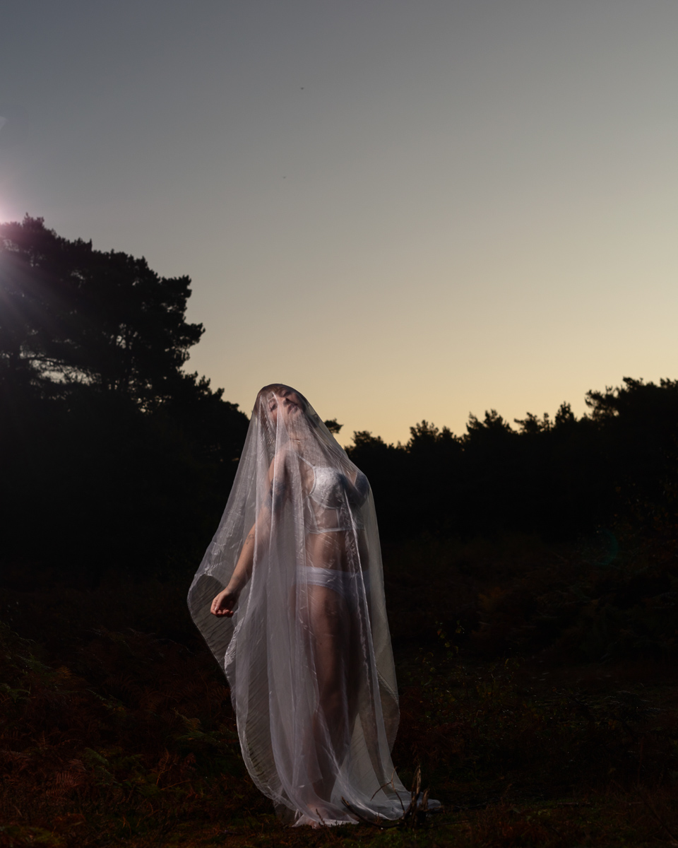 Woman draped in sheer fabric standing in forest at dusk