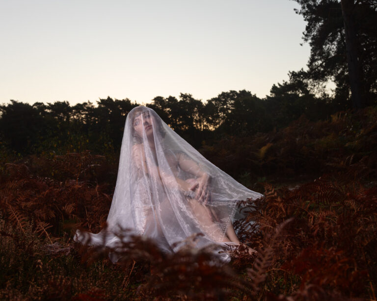 Person draped in white veil sitting in autumn fern field