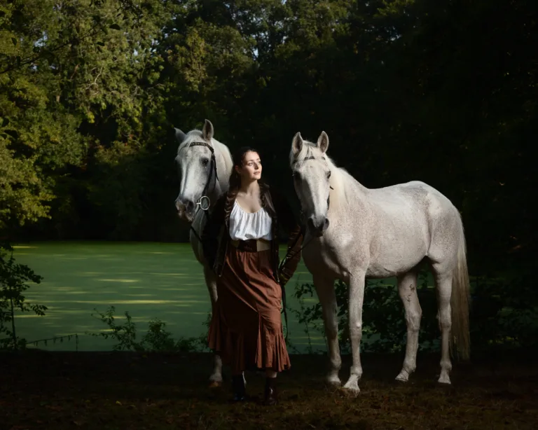 Woman in period dress standing with two white horses by water